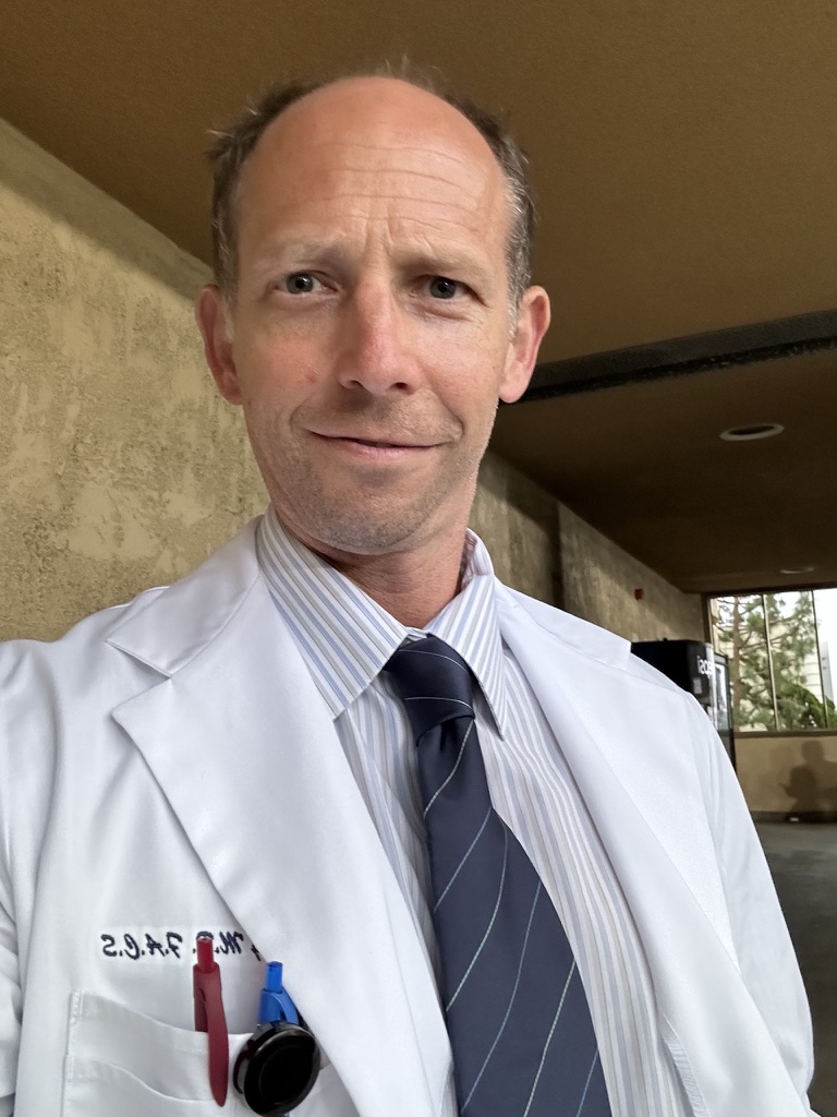 Randy Loveridge in a white doctor coat, striped shirt, and tie on the set of Grey's Anatomy